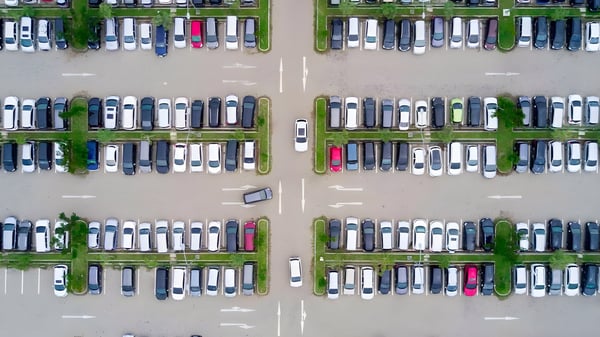 Top-down aerial view of a large outdoor parking lot with rows of cars separated by green grass medians and white arrows.