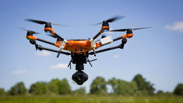 Orange hexacopter drone with a gimbal camera hovering over a green field against a blue sky.