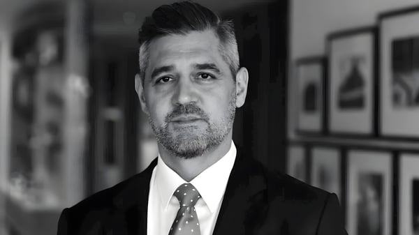 Black and white headshot of a man in a suit and patterned tie standing in front of a wall of framed pictures.