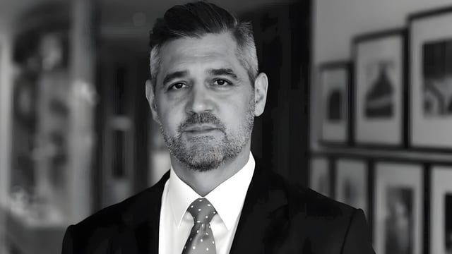 Black and white headshot of a man in a suit and patterned tie standing in front of a wall of framed pictures.