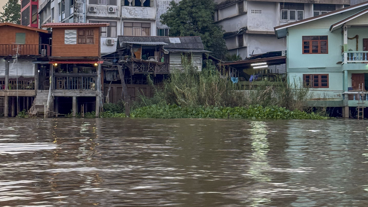 Wooden stilt houses and buildings line the edge of a wide, murky river with green vegetation growing along the bank