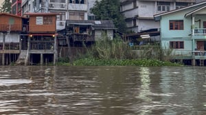 Wooden stilt houses and buildings line the edge of a wide, murky river with green vegetation growing along the bank