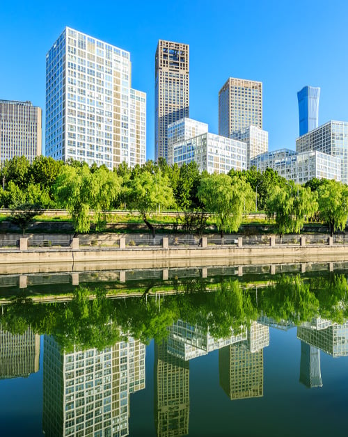 Modern city skyscrapers and green trees reflected in a calm urban river under a clear blue sky