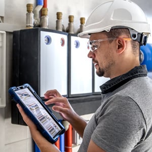 A technician in a hard hat and safety glasses uses a rugged tablet to inspect building heating and plumbing systems.