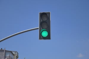 A vertical traffic light displaying a green signal against a clear blue sky, mounted on a curved metal pole.
