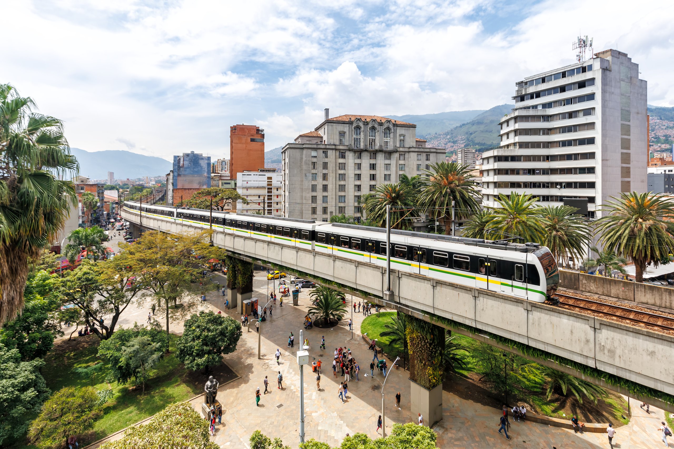 Elevated Medellin Metro train traveling on a concrete viaduct above a pedestrian plaza with palm trees and buildings