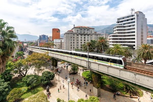 Elevated Medellin Metro train traveling on a concrete viaduct above a pedestrian plaza with palm trees and buildings