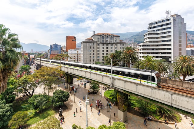 Elevated Medellin Metro train traveling on a concrete viaduct above a pedestrian plaza with palm trees and buildings