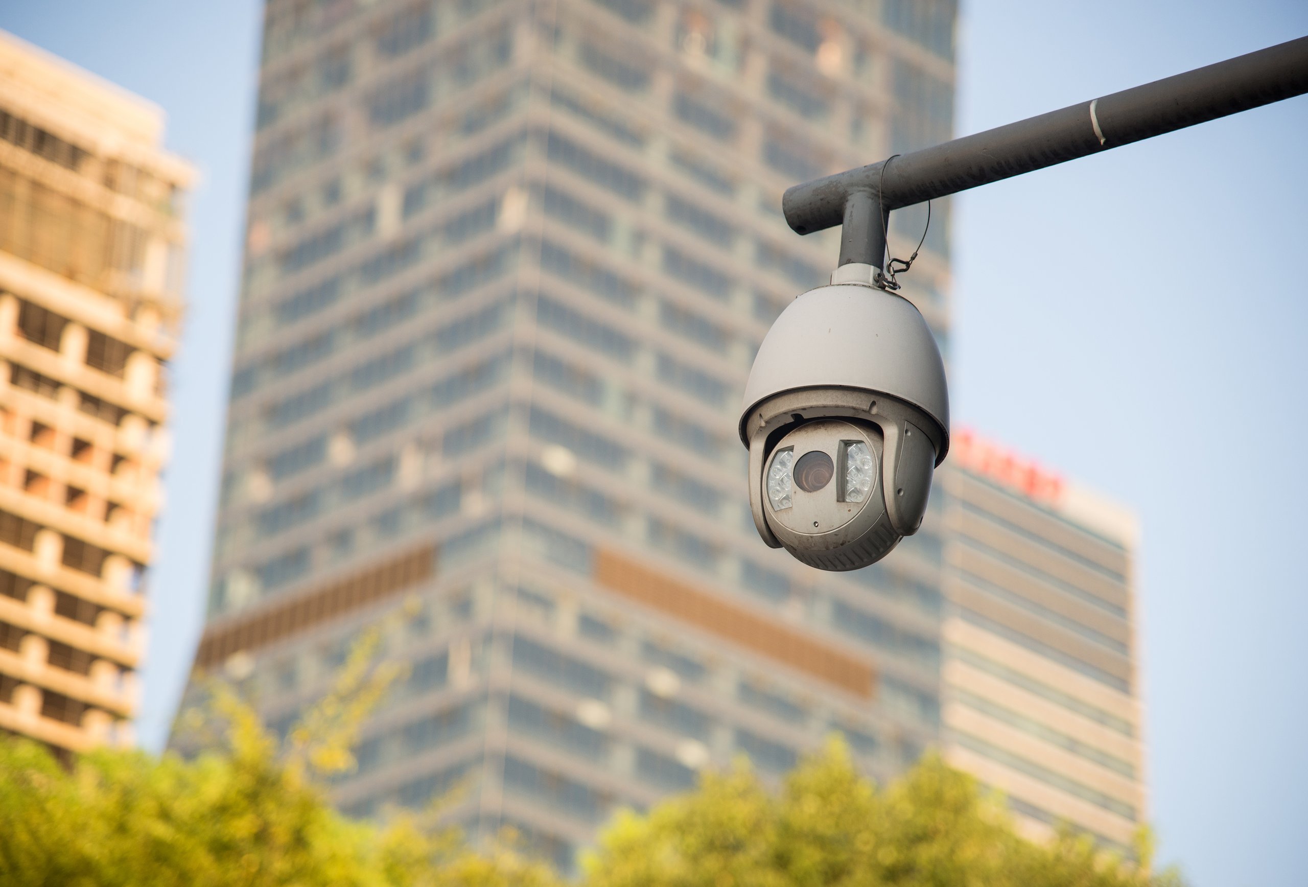 A dome security camera mounted on a metal pole with blurred city skyscrapers and green trees in the background