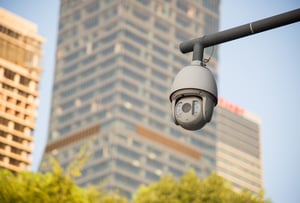 A dome security camera mounted on a metal pole with blurred city skyscrapers and green trees in the background
