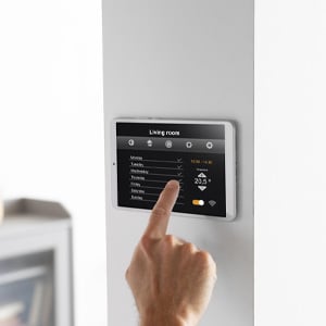 A hand adjusting the temperature on a wall-mounted smart home control panel displaying a weekly schedule.