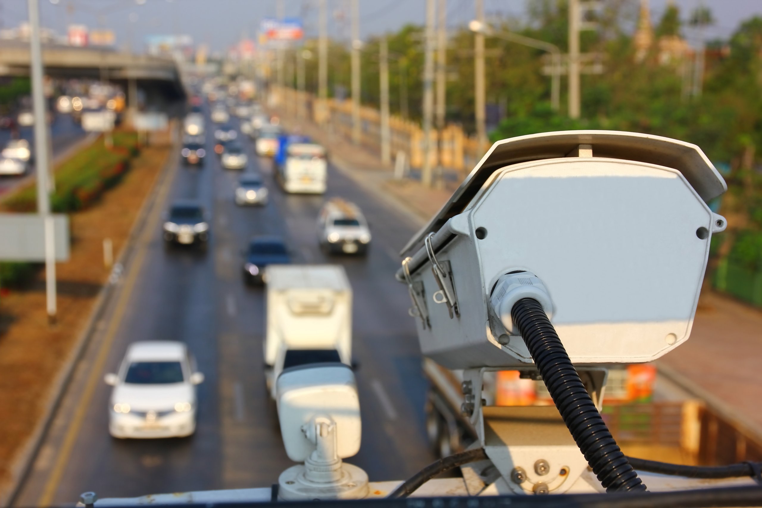 A white surveillance camera overlooks a busy multi-lane highway with moving cars and trucks.