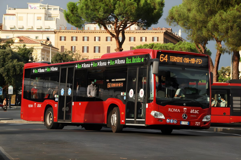 Red Roma ATAC hybrid bus driving on a city street in Rome with buildings and trees in the background.