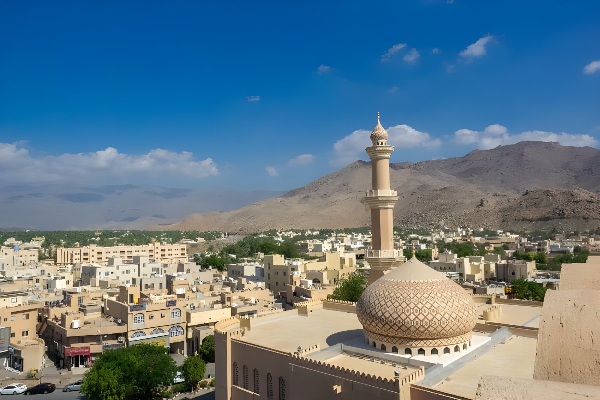 Elevated view of a mosque dome and minaret in an Omani city with arid mountains in the background.
