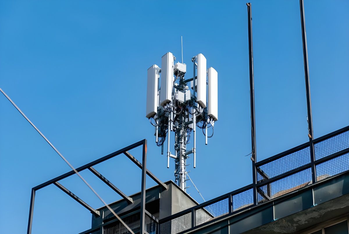 Cellular base station with white panel antennas mounted on a metal rooftop structure against a clear blue sky.