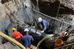 Three workers in hard hats use a chain hoist to position a large water main pipe inside a deep earthen trench.