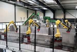 Yellow Waste Robotics arms sorting materials over a conveyor system inside a large industrial waste management facility.