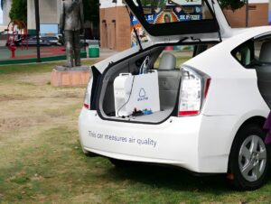 Aclima air quality monitoring equipment inside the open trunk of a white car parked near a park and statue.