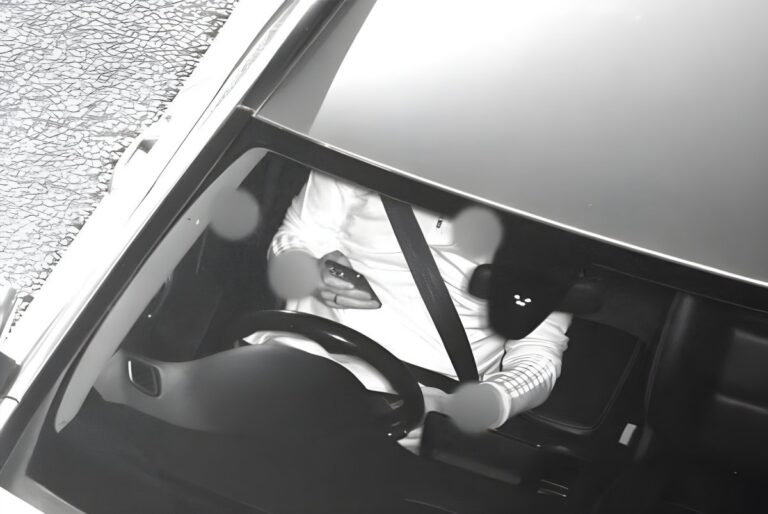 Black and white Acusensus camera view of a driver holding a mobile phone behind a steering wheel.