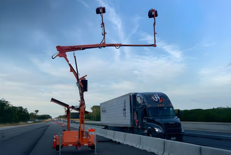 An Acusensus mobile trailer with an overhead camera arm positioned over a highway as a semi-truck drives past.