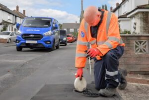 A technician in high-visibility gear opens a street utility cover near a blue Affinity Water van on a residential road.