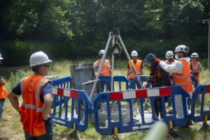 Utility workers in safety vests and hard hats use a tripod hoist at a fenced-off manhole in a grassy field.