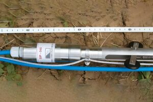 An Agisco SIP-045 geotechnical sensor probe with blue cabling and a measuring tape on dry, cracked earth.