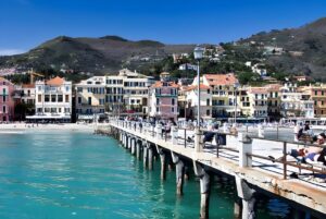 Pedestrians on the Molo di Alassio pier lined with street lamps overlooking the coastal town and hills.
