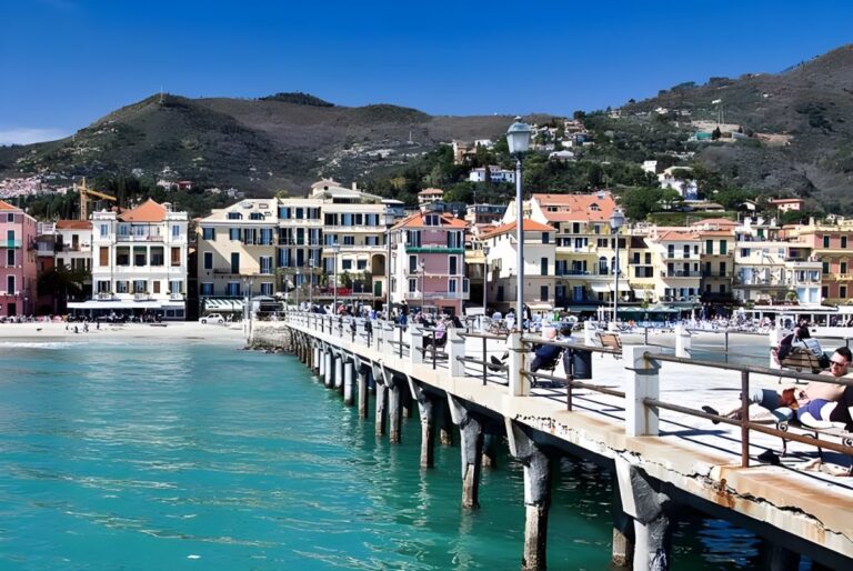 Pedestrians on the Molo di Alassio pier lined with street lamps overlooking the coastal town and hills.