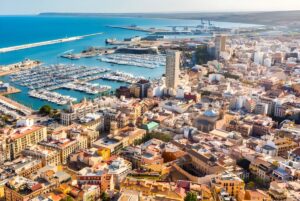 Aerial view of the Alicante coastline featuring the city harbor, moored boats, and dense urban infrastructure.