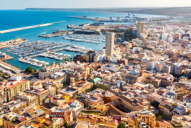 Aerial view of the Alicante coastline featuring the city harbor, moored boats, and dense urban infrastructure.