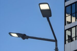Two illuminated LED cobra head streetlights on a dual-arm pole against a blue sky and office building.
