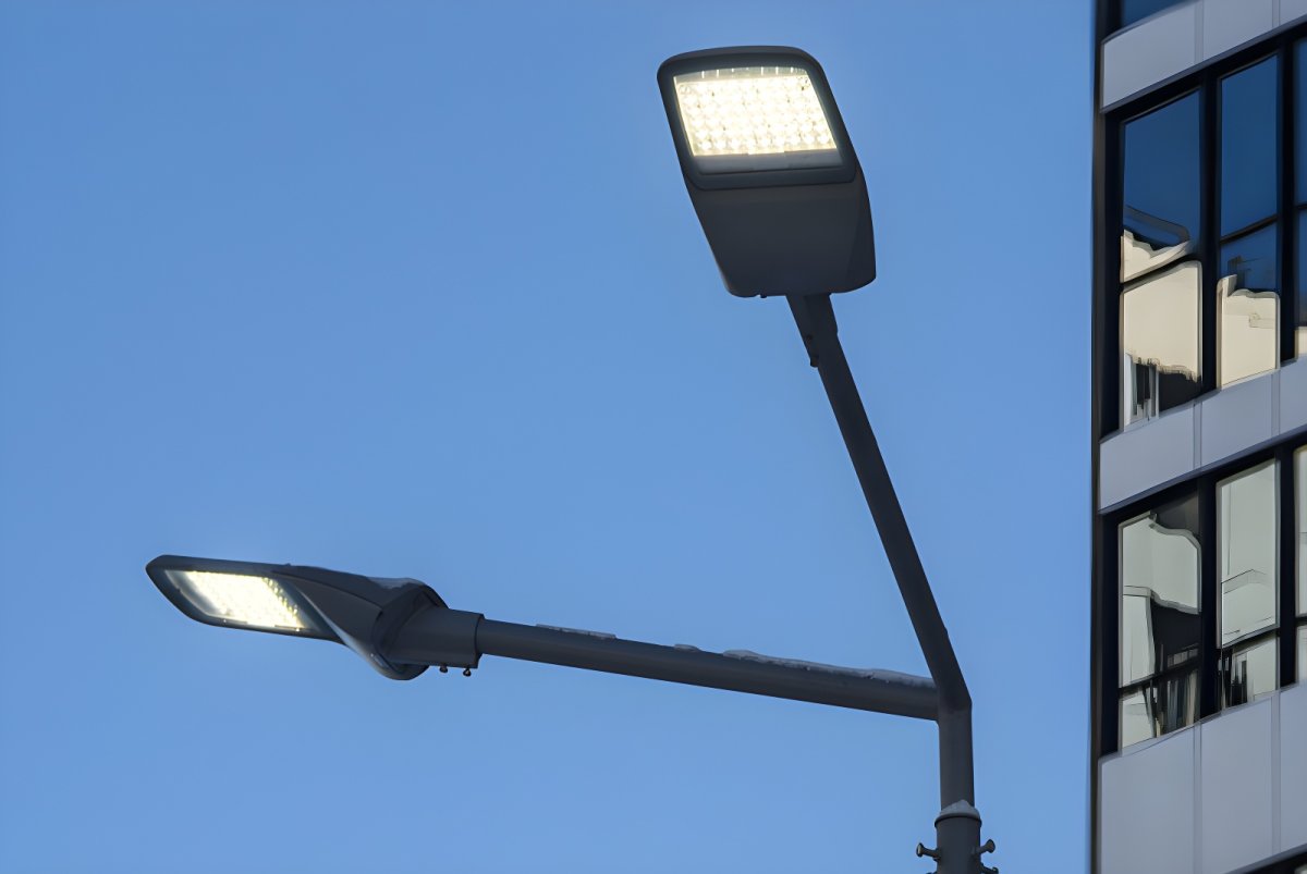 Two illuminated LED cobra head streetlights on a dual-arm pole against a blue sky and office building.