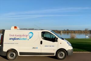 White Anglian Water service van parked on a paved road beside a lake under a clear blue sky.