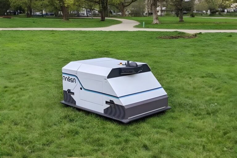 An Angsa Robotics autonomous cleaning robot on a green lawn in a public park with trees and a walking path.