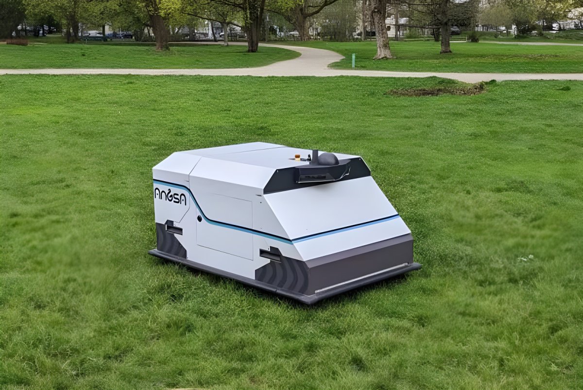 An Angsa Robotics autonomous cleaning robot on a green lawn in a public park with trees and a walking path.