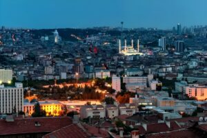 High-angle evening cityscape of Ankara, Türkiye, featuring illuminated buildings and the Kocatepe Mosque in the distance.