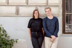 Another Earth founders standing against a stone wall with a window and green foliage.