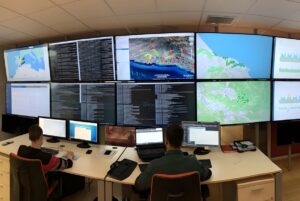 Two operators at a desk monitoring a large wall of screens displaying digital maps, data code, and utility analytics.