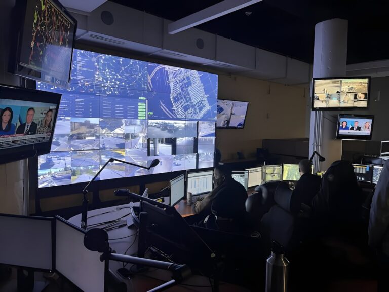 Operators in a real-time crime center monitor a large video wall displaying city maps and live surveillance camera feeds.