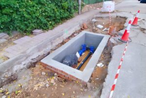 Concrete utility vault under construction on a roadside featuring blue water valves, pipes, and orange traffic cones.