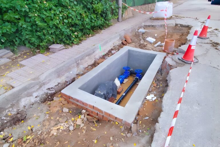 Concrete utility vault under construction on a roadside featuring blue water valves, pipes, and orange traffic cones.