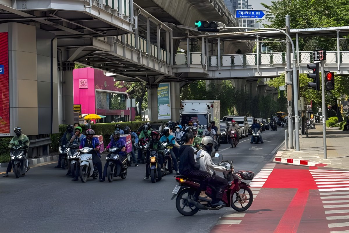 Motorcyclists wait at a Bangkok intersection under a pedestrian skywalk with active traffic signals and red crosswalks.