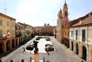 High-angle view of a historic Italian town square with parked cars, arched walkways, and a twin-towered church.