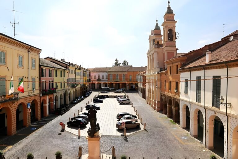 High-angle view of a historic Italian town square with parked cars, arched walkways, and a twin-towered church.