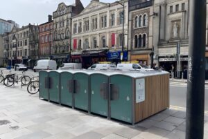 A row of green and wood-paneled BikeDok solar-powered cycle lockers on a paved city sidewalk in front of historic buildings.