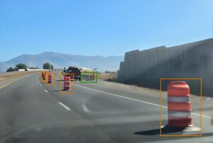 Dashcam view of a highway with AI bounding boxes identifying orange traffic barrels and a construction barrier.