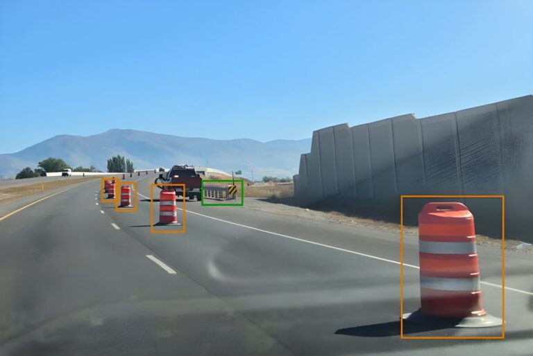 Dashcam view of a highway with AI bounding boxes identifying orange traffic barrels and a construction barrier.