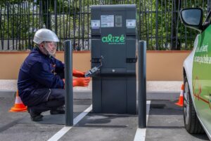 A technician in a hard hat and face shield services an Alizé EV charging station next to a parked car.