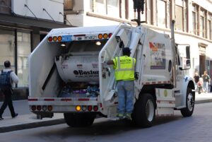 A sanitation worker in a neon vest rides on the back of a white Boston Public Works garbage truck on a city street.